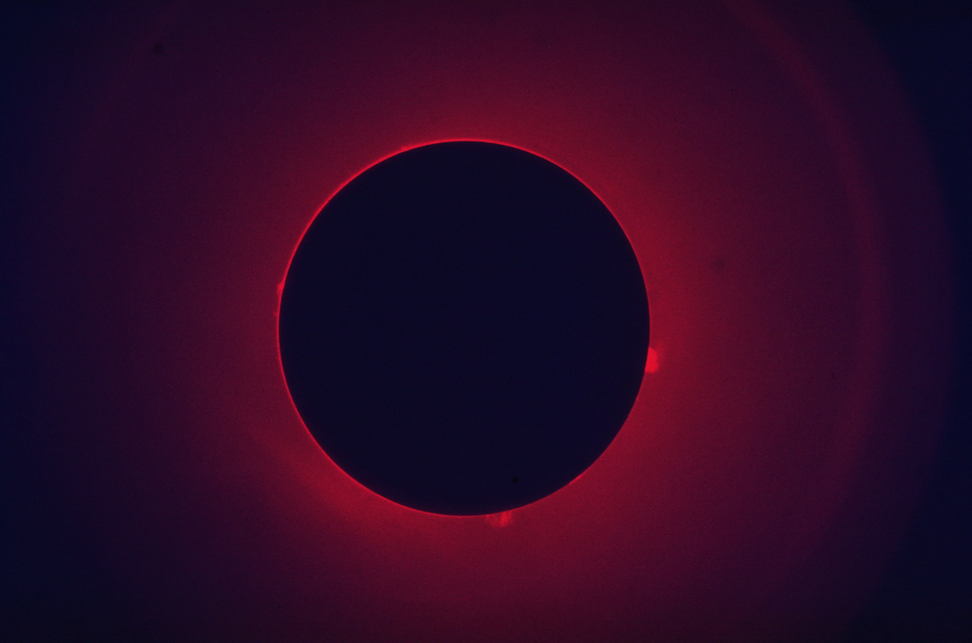 Vedyn alfaviivalla kuvattu Aurinko, jonka tumman kiekon ympärillä näkyy protuberansseja.